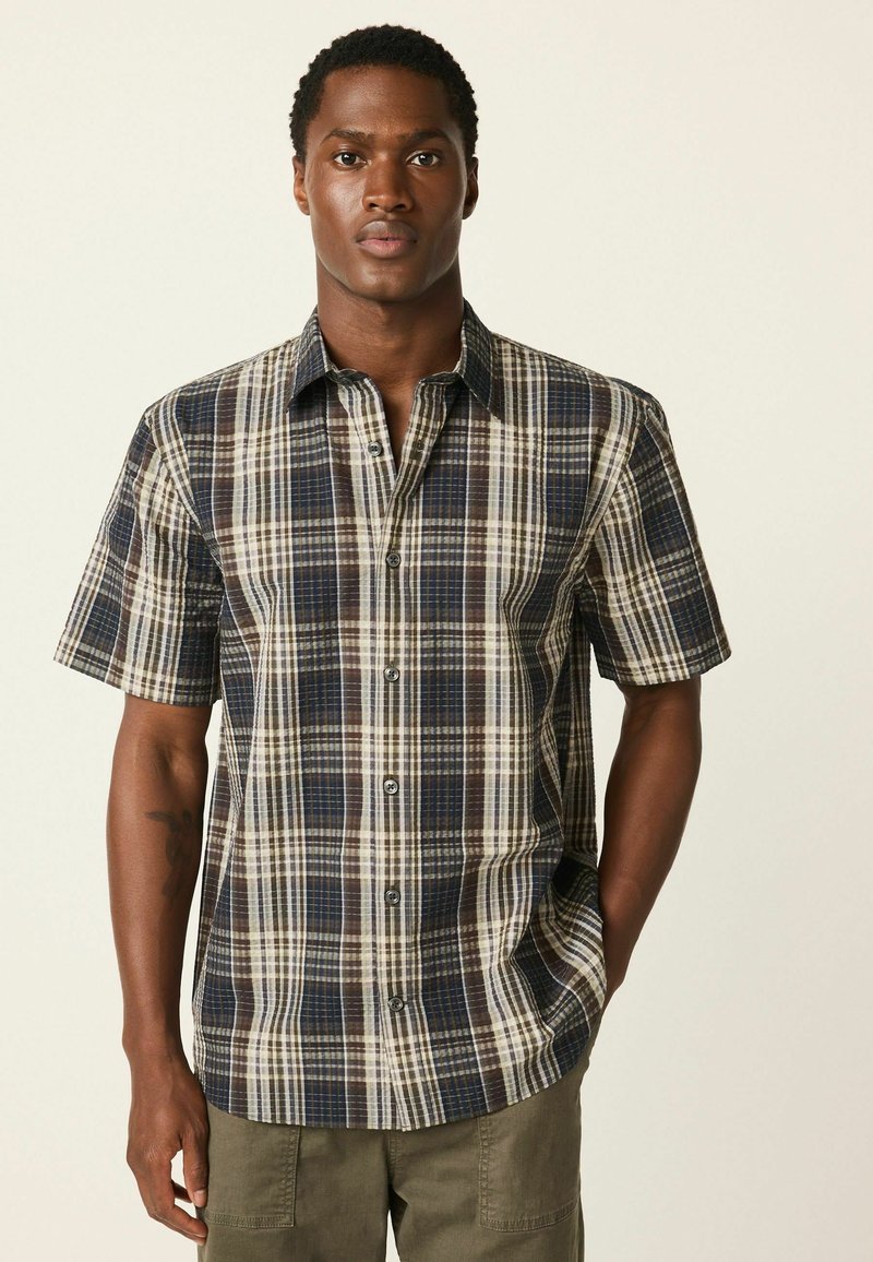 Košeľa s krátkym rukávom s károvaným vzorom v námorníckej modrej, hnedej a krémovej farbe. Má zapínanie na gombíky vpredu a pohodlný strih s jemným leskom v textúre látky.