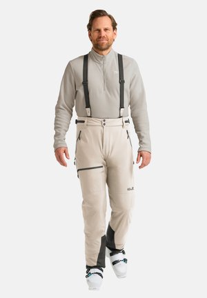Beige Skihosen mit schwarzen Akzenten, verstellbaren Hosenträgern und Reißverschlusstaschen, kombiniert mit einem hellgrauen Langarmshirt und Skistiefeln.
