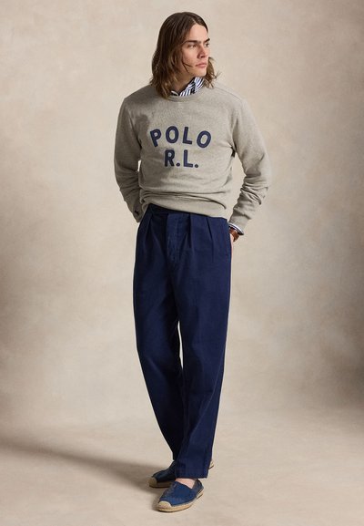 Homme portant un sweat-shirt gris avec le texte « POLO R.L. », une chemise rayée, un pantalon plissé bleu marine et des espadrilles bleues, debout les mains dans le dos.