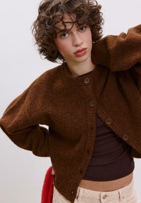 Hnědý pletený cardigan s kulatým výstřihem, předními knoflíky a žebrovanými manžetami. Model má pod sebou přiléhavé tmavé tričko.