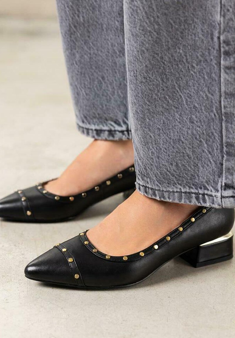 Zapatos de cuero negro con punta afilada, adornados con acentos de tachuelas doradas y un tacón bajo y cuadrado. El diseño presenta una textura suave y bordes curvados.