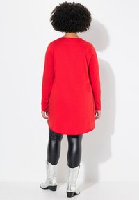 Rote Tunika mit langen Ärmeln und abgerundetem Saum, kombiniert mit schwarzen Leggings und silbernen Cowboy-Stiefeln. Glattes Material, legere Passform.