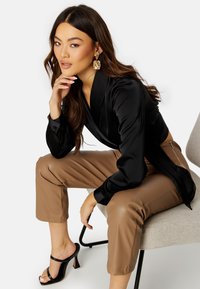 Blusa avvolgente in raso nero, pantaloni fitted beige e tacchi a piedi aperti neri. Modella seduta su una sedia elegante, che mette in mostra texture e colori raffinati.