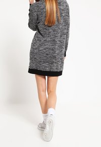 Graues Strickkleid mit schwarzem Saum, langen Ärmeln und weicher Textur. Getragen mit weißen Socken und grauen Sneakers, die Rückansicht wird gezeigt.
