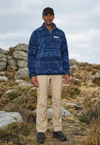 Man die een blauwe fleece met patroon draagt, beige cargobroek en een zwarte pet en schoenen, staande op een rotsachtig, grasachtig wandelpad.