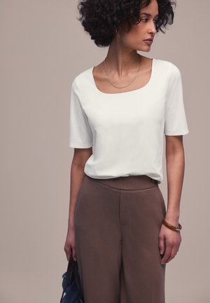 Frau mit kurzen, lockigen Haaren trägt ein weißes Oberteil mit quadratischem Ausschnitt, braune hochgeschnittene Hose, goldene Halskette und brauen Armreif und hält eine blaue Tasche.