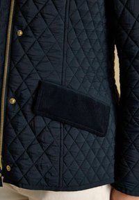 Jachetă quilted de culoare marină, cu un fermoar auriu, model în diamant și detalii din catifea. Textură moale cu un finisaj neted.