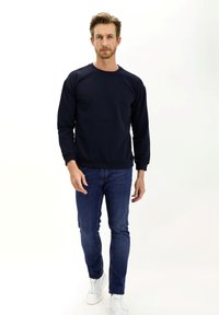 Sudadera azul marino de material polar con puños y dobladillo acanalados, combinada con jeans de mezclilla oscuros y zapatillas blancas. Diseño minimalista.
