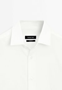 Chemise blanche boutonnée avec un col pointu et des étiquettes noires indiquant la marque et l'origine de fabrication à l'intérieur du col.
