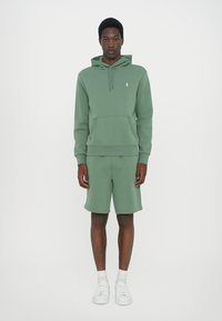 Sweatshirt verde de algodão e calções coordenados, apresentando um bolso canguru, punhos canelados e detalhe em logo branco. Combinado com ténis brancos.