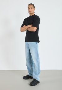 T-shirt noir à manches courtes avec détail de logo, jeans bleus clair à coupe décontractée, et chaussures noires à semelles épaisses. Tenue simple et décontractée.
