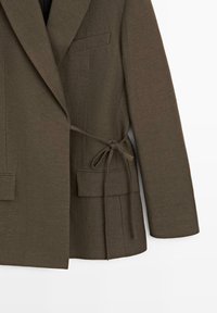 Veste marron sur mesure avec col à revers, deux poches à rabat, une poche poitrine et une ceinture en tissu nouée sur le côté droit.