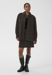 Brun ruskindsjakke med forlommer, båret over en sort top og matchende nederdel. Sorte knæhøje støvler fuldender outfittet.