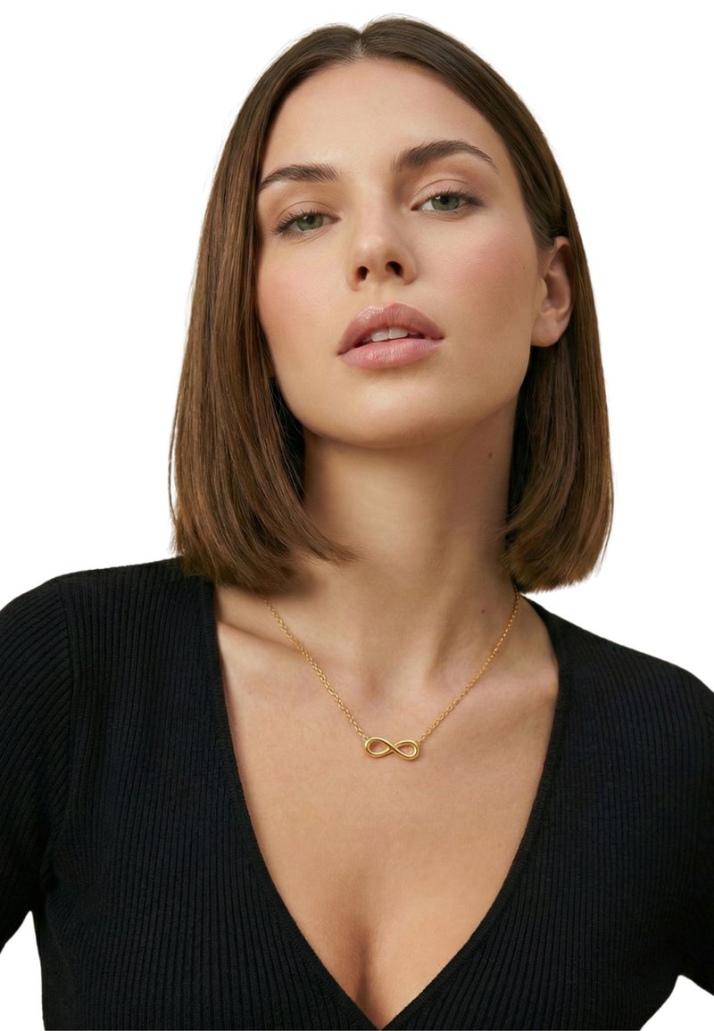 Jeune femme aux cheveux bruns raides, portant un haut noir à col en V et un collier avec un symbole d'infini en or, regardant légèrement vers le haut.