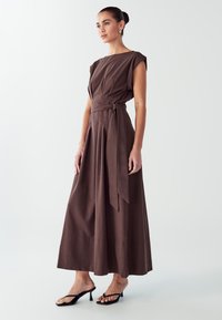 Hnědé maxi šaty bez rukávů s přitaženým pasem a detailem na zavazování. Hladká textura, silueta ve tvaru A a jemné průhyby podél živůtku.