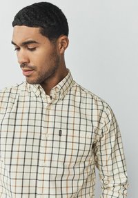 Camisa de botones con una base crema que presenta un patrón de cuadros multicolores en negro, marrón y verde. Bolsillo en el lado izquierdo del pecho con detalle de logo.