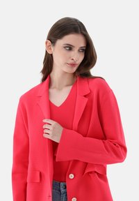 Roter Blazer mit Reverskragen, langen Ärmeln, frontalen Knöpfen und Seitentaschen, über einem roten gerippten Oberteil getragen. Glatte Textur, taillierter Schnitt.