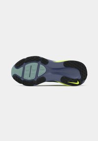 Sportinių batų padas su juodais, mėlynais ir neoniniais geltonais gumos sukibimais, šalia kulno matomas mažas geltonas Nike swoosh.