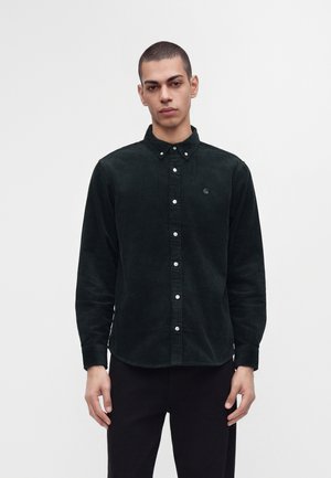 Jeune homme aux cheveux courts portant une chemise en velours côtelé vert foncé boutonnée et un pantalon noir, debout devant un fond blanc uni.