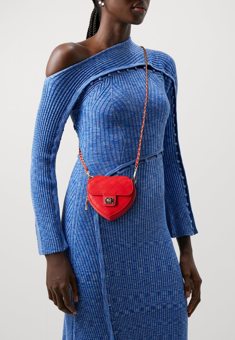 Bolso rojo acolchado en forma de corazón con cadena. Llevado sobre un vestido azul texturizado de hombros al descubierto, con tela acanalada y detalles de botones.