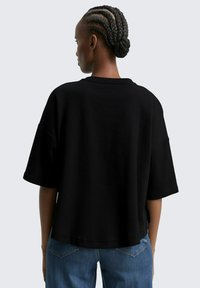 T-shirt noir oversize, à manches courtes, texture côtelée, col rond. Porté avec un jean en denim bleu, offrant une coupe décontractée et des poches arrière.