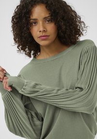 Pulover verde deschis dintr-un material moale, cu mâneci lungi plisate și croială lejeră. Guler rotund și detalii cu tiv elastic.