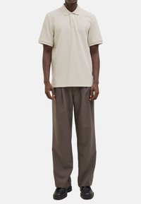 Beige strukturierte Polo-Shirt mit Kragen und drei Knöpfen, kombiniert mit locker sitzenden dunkelbraunen Hosen und schwarzen Schuhen.