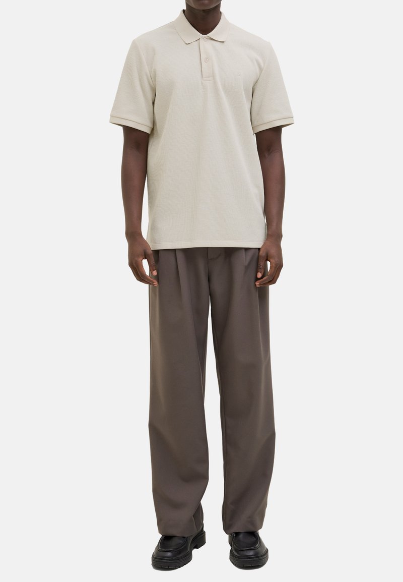 Beige strukturierte Polo-Shirt mit Kragen und drei Knöpfen, kombiniert mit locker sitzenden dunkelbraunen Hosen und schwarzen Schuhen.