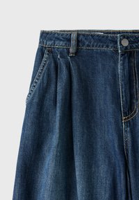 Jupe en denim bleu foncé avec plis à l'avant, poches latérales, passants de ceinture et fermeture par bouton métallique sur fond gris clair.