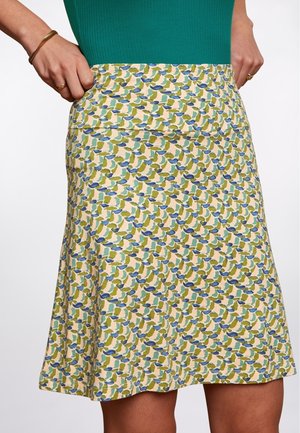 Kobieta ubrana w spódnicę o kroju litery A sięgającą do kolan, z abstrakcyjnym wzorem w odcieniach niebieskiego, zielonego i beżowego, w połączeniu z zielonym topem bez rękawów.