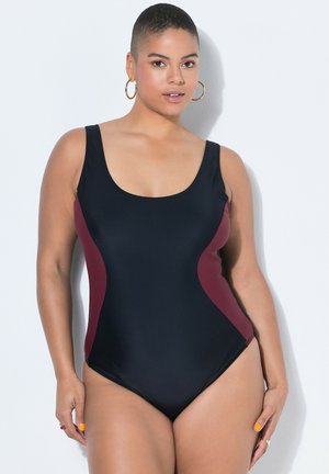 Mujer con un traje de baño de una pieza negro con paneles laterales burdeos, de pie contra una pared blanca, con pendientes de aro y anillos.