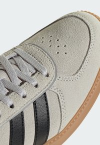 Svjetlosiva platnena tenisica sa crnim kožnim prugama, teksturiranim gornjim dijelom, perforiranim detaljem na vrhu i smeđom gumom na potplatu.