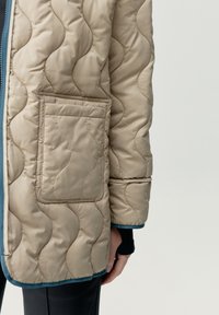 Giacca trapuntata beige con motivo a onde, dotata di tasca a toppa e rifiniture color teal all'orlo e ai polsini, realizzata in tessuto leggero e lucido.