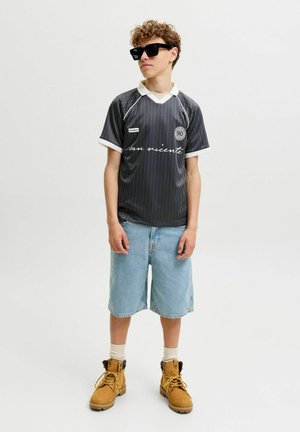 Persona joven con cabello rizado, que lleva gafas de sol negras, camisa oscura de rayas con mangas cortas, pantalones cortos de mezclilla claros, calcetines blancos y botas marrones.