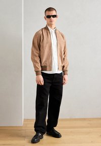 Braune Wildleder-Bomberjacke mit grünen und roten Rippakzenten, weißem Hemd, schwarzen Hosen und schwarzen Schuhen.
