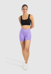 Schwarzes, kurzes Tanktop kombiniert mit hoch geschnittenen lila Shorts. Glattes Material, eng anliegendes Design und weiße Sportschuhe.