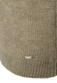 Nahaufnahme von beigem Strickstoff mit geripptem Saum und einem kleinen rechteckigen metallischen Markenetikett, das nahe der unteren Kante angebracht ist.