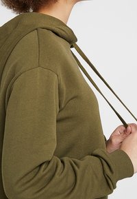 Sudadera con capucha de color verde oliva con cordón, hecha de tela suave. Cuenta con un ajuste relajado y mangas raglán para facilitar el movimiento.