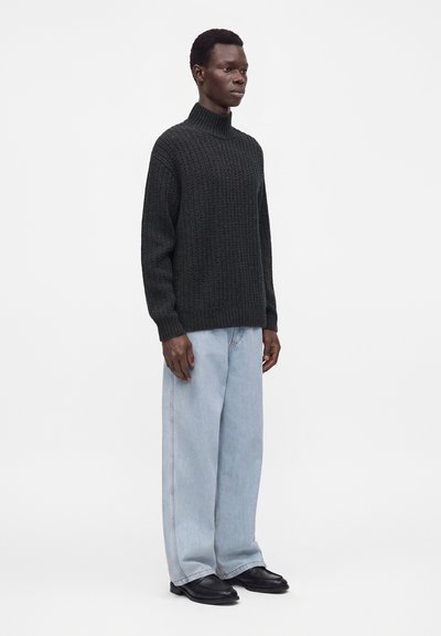 Homme debout portant un pull à col roulé texturé foncé, un jean large bleu clair et des chaussures noires sur fond blanc uni.