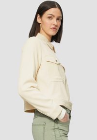 Korte denimjas in lichtbeige met knoopsluiting aan de voorkant, twee borstzakken en geribbelde manchetten. Gemonteerd met olijfgroene broek.