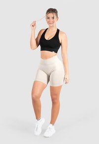 Zwarte sportbeha en beige shorts met hoge taille gedragen met witte sneakers. Glad, aansluitend materiaal met een strak ontwerp. Neutrale achtergrond.