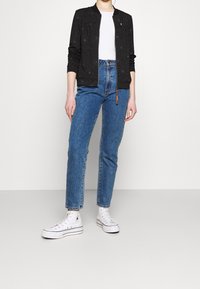 Svart bomberjacka med små vita prickar, ihopparad med högmidjade blå jeans och vita kanvas sneakers. Enkel krage och dragkedjestängning.