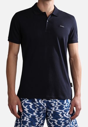Navy polo shirt in soft cotton fabric with a classic collar, three buttons, and a branded tag on the hem. Paired with patterned swim trunks.