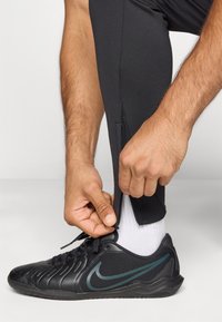 Melnas sporta bikses ar rāvējslēdzēja sānu, pieguļošs dizains; cilvēks pielāgo uzmavu. Melnas un zaļas Nike apavi, redzami balti zeķes.