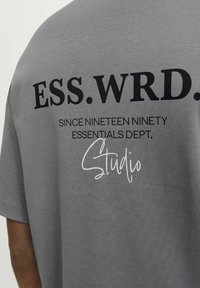 Grå bomulds-t-shirt med korte ærmer, der har bold sort tekst: "ESS. WRD." og "SINCE NINETEEN NINETY ESSENTIALS DEPT." i hvid skrift.