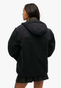 Zwarte fleece jacket met een gestructureerde oppervlakte, met een capuchon en een contrasterend zwart bovenstuk. De jacket is gedragen over een geruite rok.