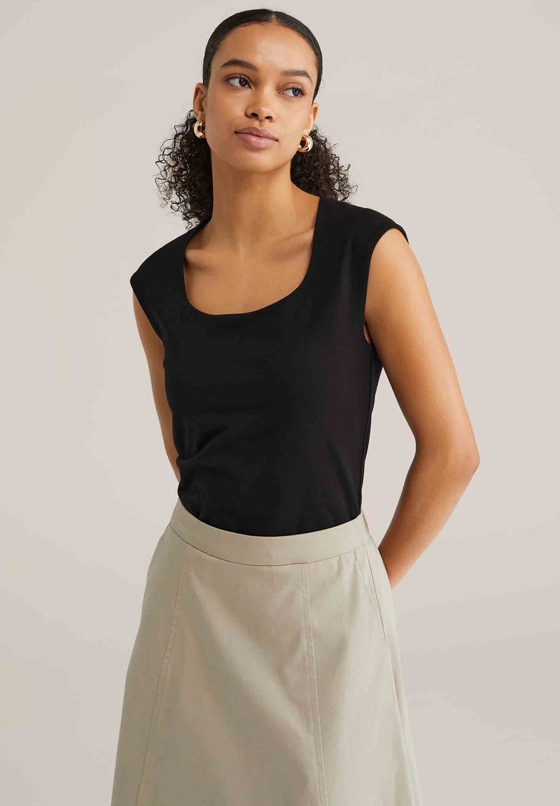 Jeune femme aux cheveux bouclés portant des boucles d'oreilles créoles en or, un haut noir sans manches et une jupe beige, debout les mains derrière le dos.