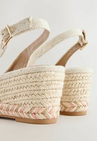 Bézs színű, fonott ékített szandálok bokapántokkal és arany csatokkal, fonott kötél sarokkal, rózsaszín és fehér részletekkel.