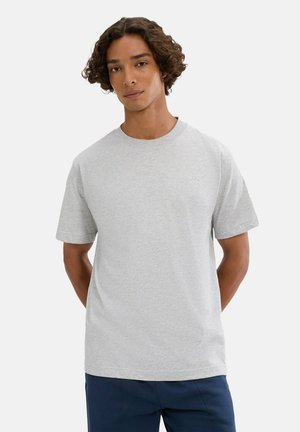 Joven con pelo rizado que lleva una camiseta de cuello redondo, lisa, de color gris claro y de manga corta, y pantalones oscuros, de pie con las manos detrás de la espalda.