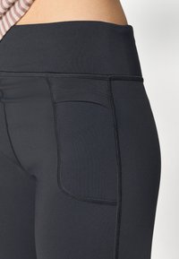 Leggings noirs en matériau lisse et extensible avec une large taille, comportant une petite poche latérale et des coutures contrastées.